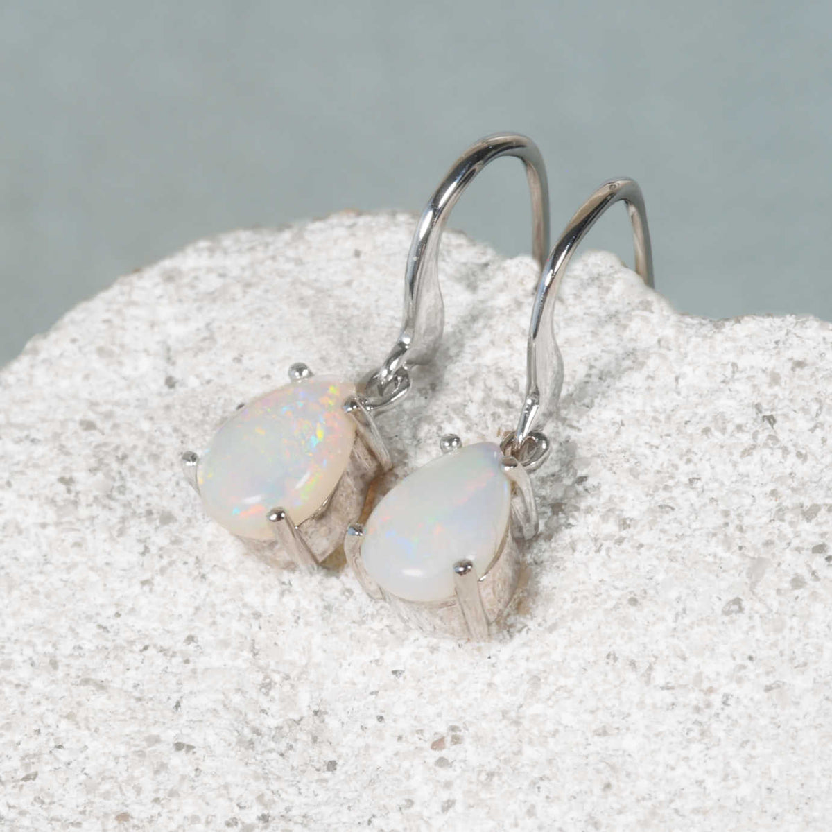 A pair of white gold teardrop-shape authentic Australian opal dangle earrings resting on a textured white stone surface against a soft gray background.