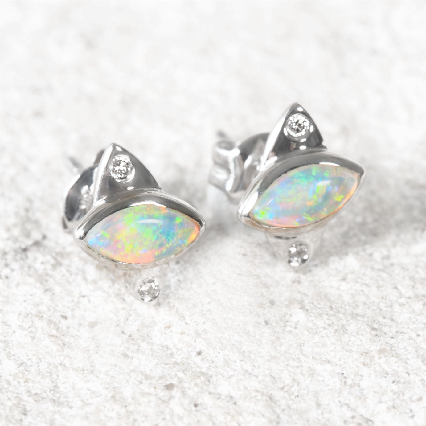 A pair of white gold opal stud earrings featuring marquise-cut iridescent Australian opal stones and small clear accent stones, displayed on a light textured surface.