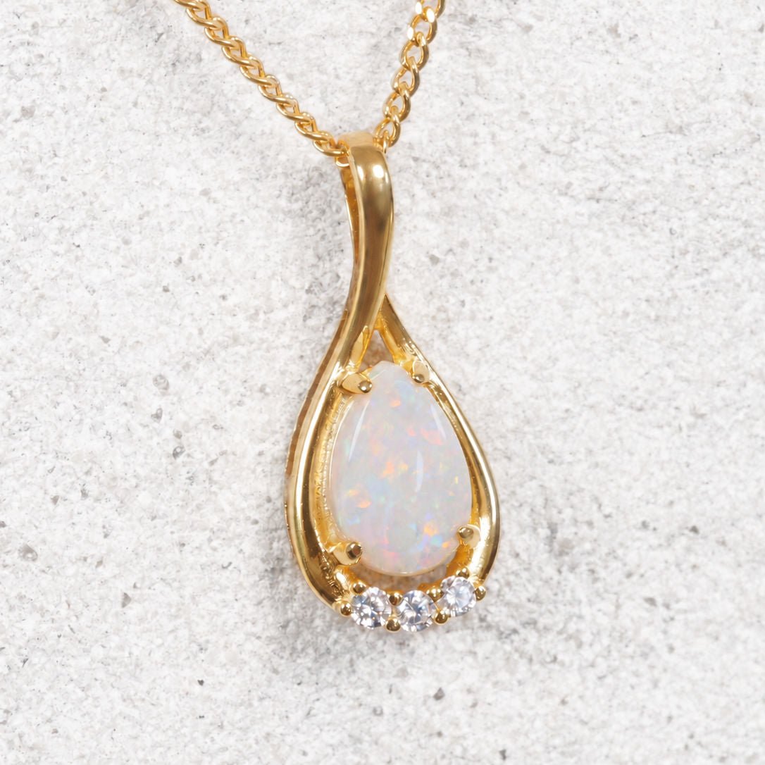 A gold plated silver teardrop pendant necklace featuring a white opal center stone and three small clear accent stones, displayed on a textured light gray surface.