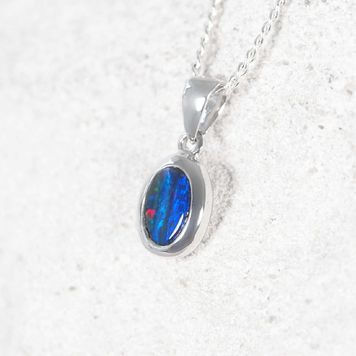 A 14ct white gold oval pendant necklace featuring a vibrant but petite blue and red opal gemstone, displayed on a textured white surface.
