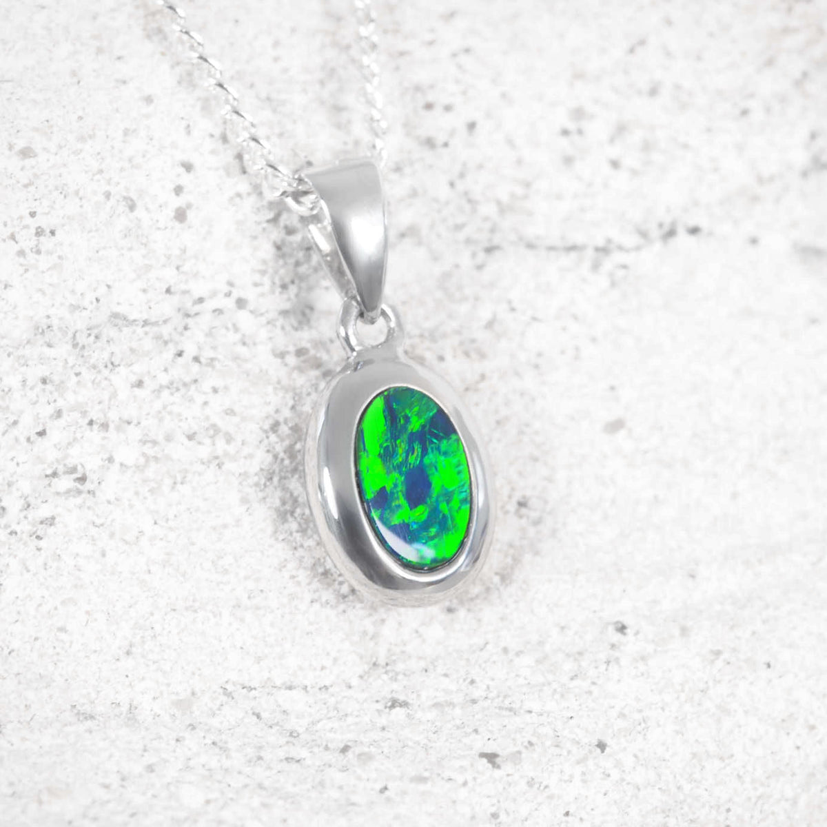 A 14ct white gold oval pendant necklace featuring a vibrant green and blue iridescent opal stone, shown on a textured light gray background.
