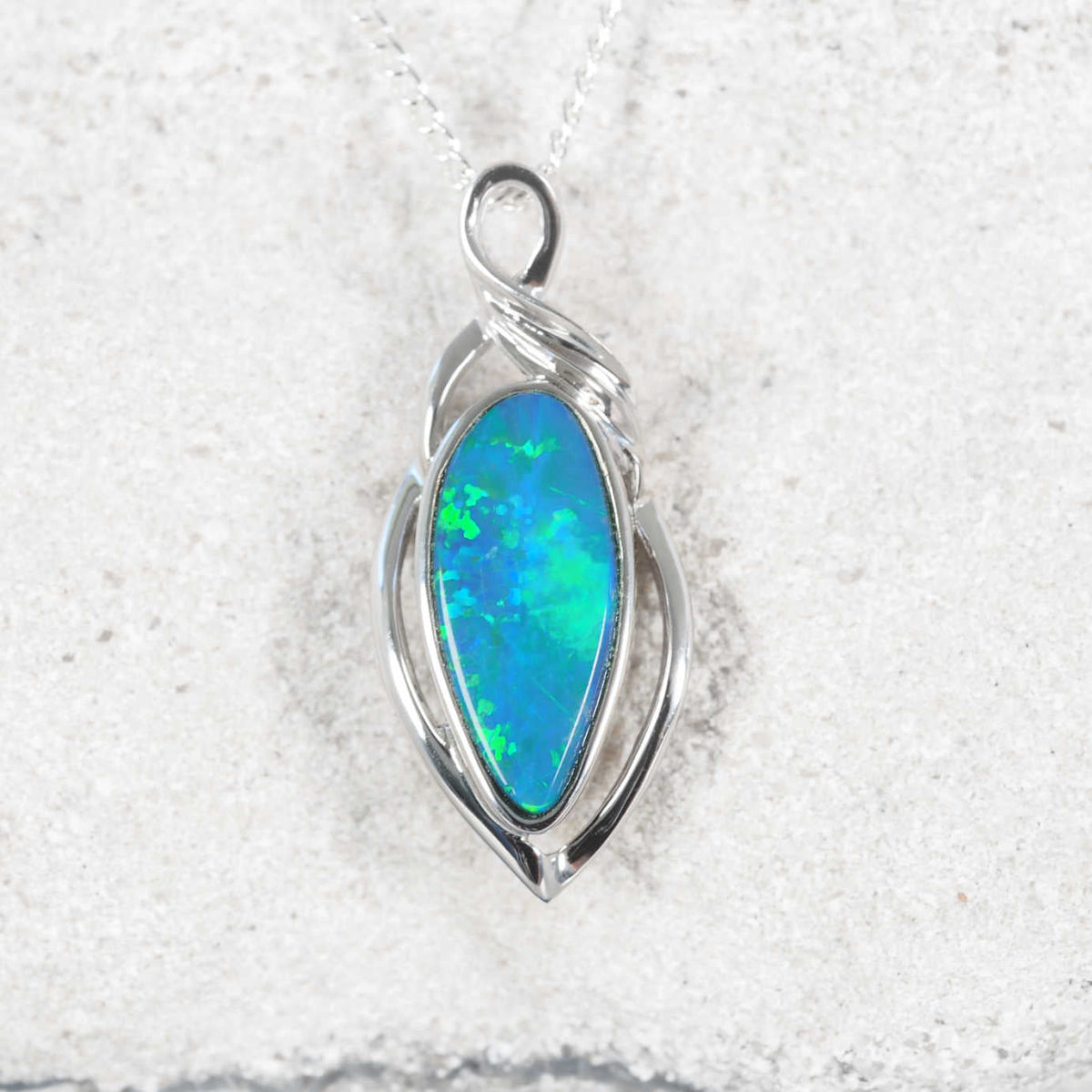 A silver opal pendant necklace featuring a teardrop-shaped blue opal gemstone, displayed on a textured light gray stone surface.