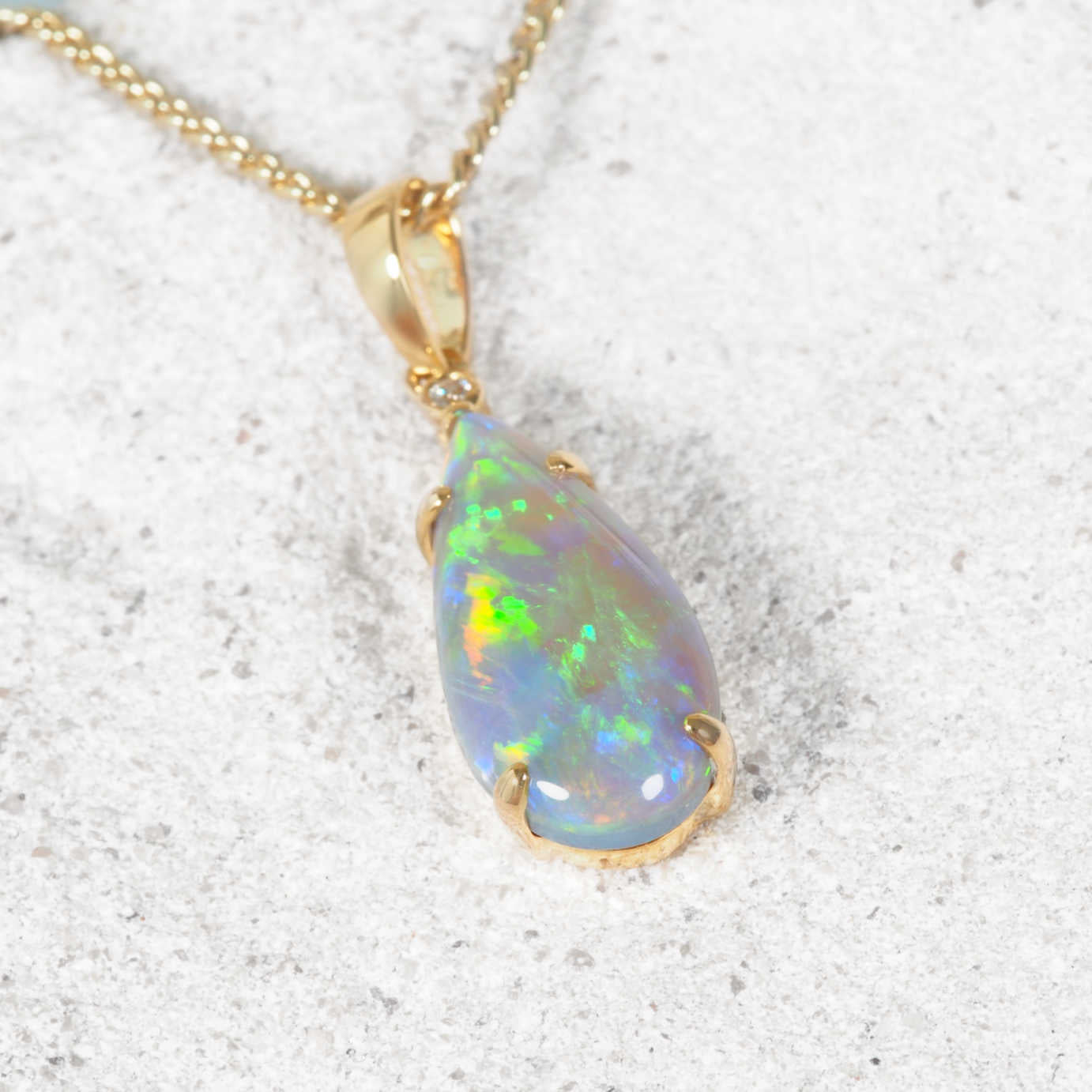 A gold teardrop pendant necklace featuring a vibrant, iridescent opal gemstone, resting on a textured light gray stone surface.