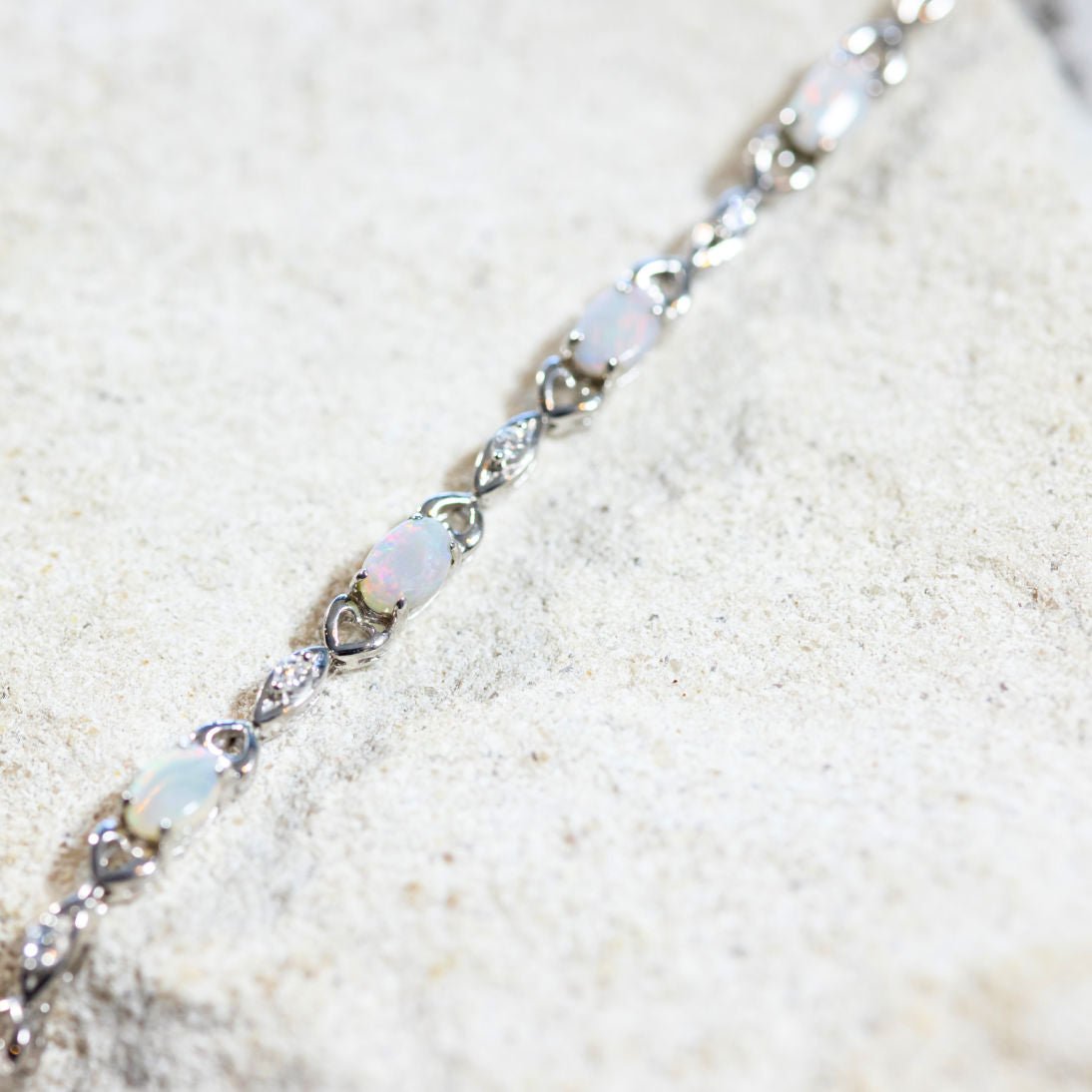 A close-up image of a sterling silver bracelet featuring a heart design with white opal gemstones and sparkling diamantés set against a neutral background.