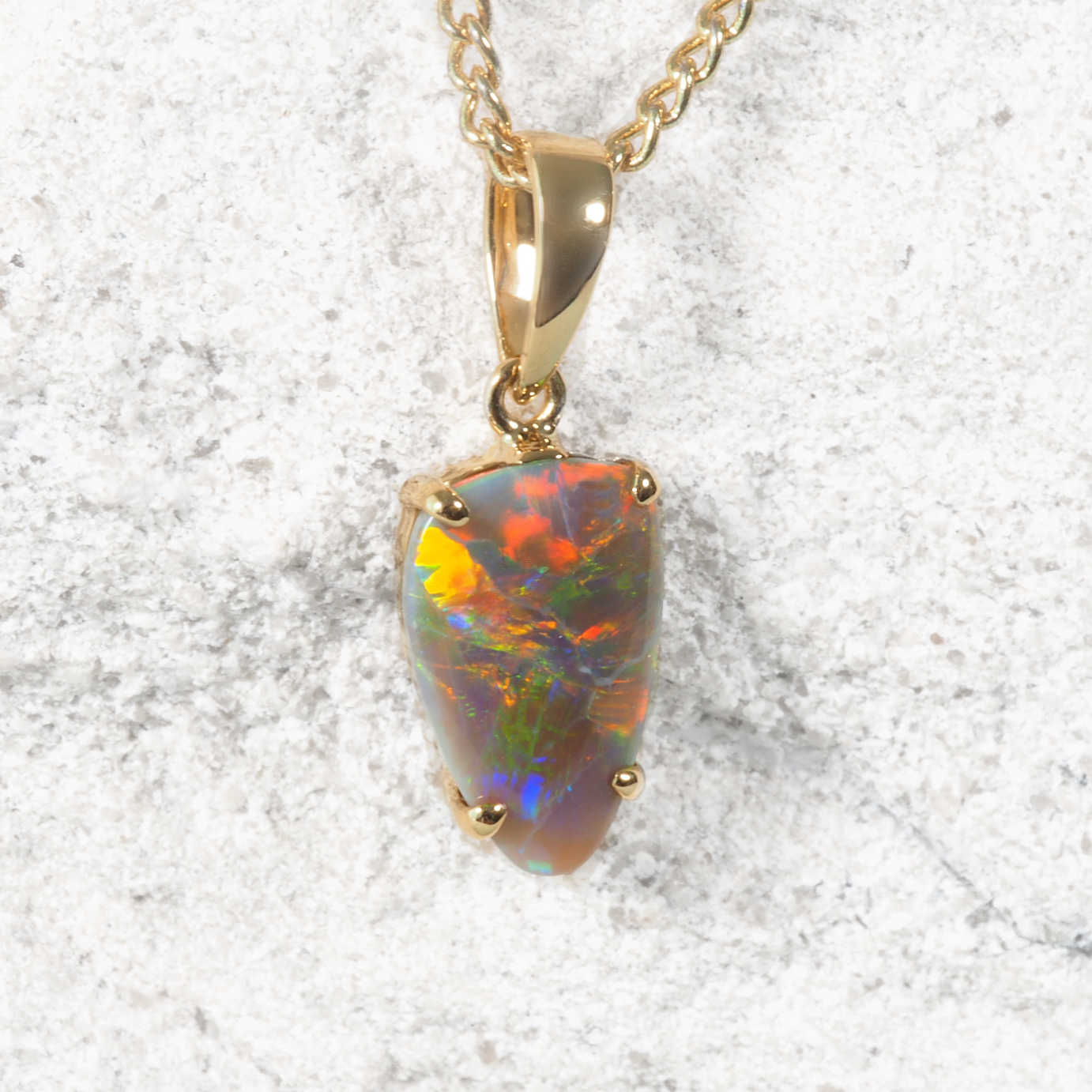 A gold authentic Australian opal necklace pendant featuring a teardrop-shaped Lightning Ridge opal stone with vibrant multicolor play-of-color, displayed on a textured light stone background.