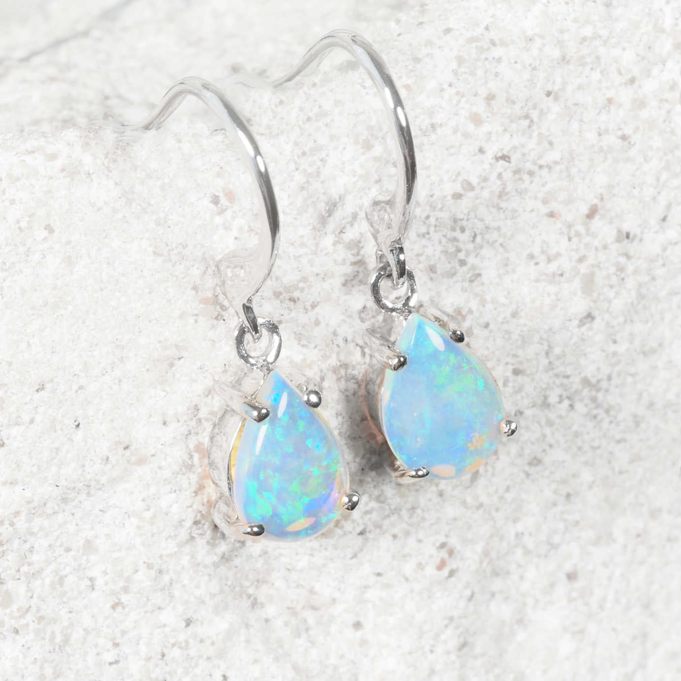 A pair of white gold opal drop earrings with teardrop-shaped blue opal gemstones, displayed on a textured light gray stone surface.