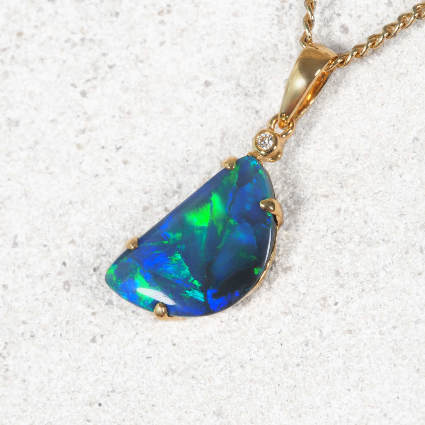 A gold pendant necklace featuring a large, irregularly shaped opal gemstone with vibrant blue and green flashes, set on a textured light gray background.
