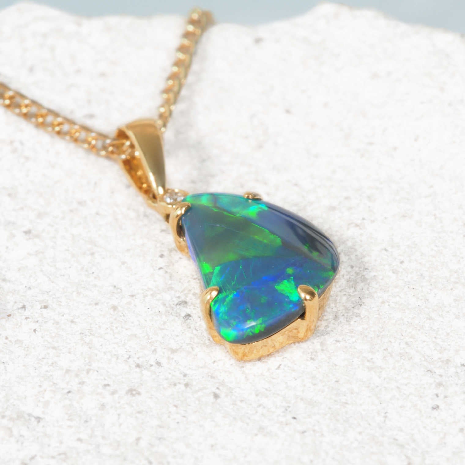 A Lightning Ridge opal gold pendant necklace featuring a teardrop-shaped iridescent opal gemstone with blue and green flashes, resting on a textured white stone surface.