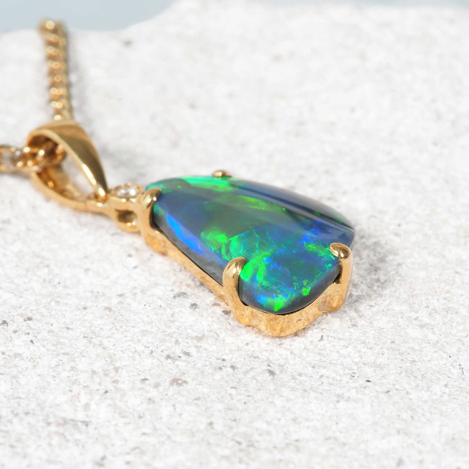 A black opal gold pendant necklace featuring a large, vibrant iridescent opal stone with blue, green, and orange flashes, resting on a textured light gray stone surface.