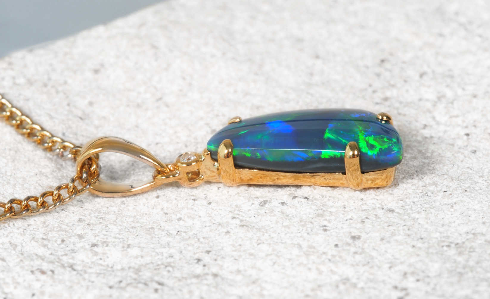 A black opal gold pendant necklace featuring a rectangular cabochon opal gemstone with vibrant blue, green, and orange play-of-color, resting on a textured light gray stone surface.