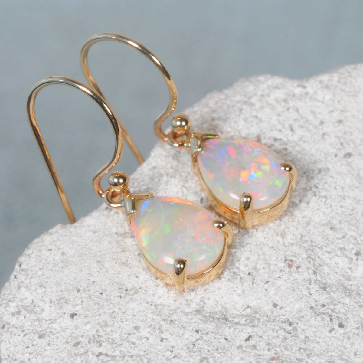 A pair of Australian opal gold drop earrings with teardrop-shaped iridescent opal stones, displayed on a textured white stone surface against a soft gray background.