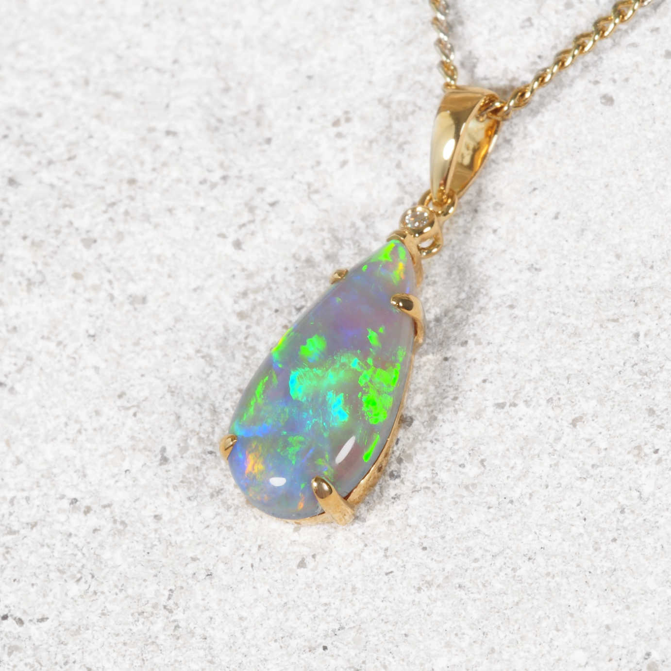 A gold teardrop pendant necklace featuring a vibrant, iridescent Australian black opal with green and blue flashes, resting on a textured stone surface.