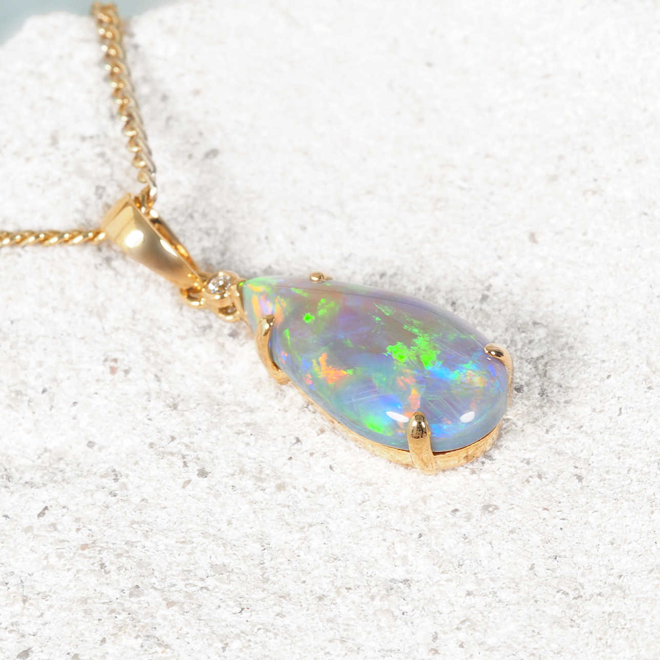 A gold teardrop pendant necklace featuring a vibrant, iridescent opal gemstone with flashes of green, blue, and orange, resting on a textured light gray stone surface.