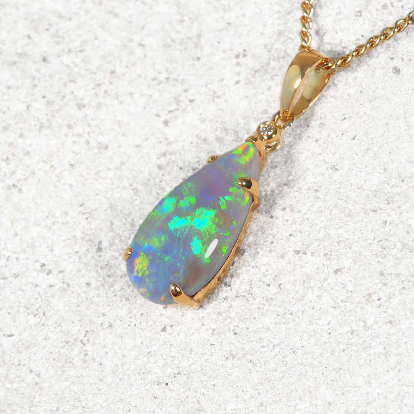 A teardrop-shaped opal pendant necklace in a gold setting on a textured light gray background. The opal displays vibrant flashes of green, blue, and white, set in a gold bezel with a small clear accent stone.