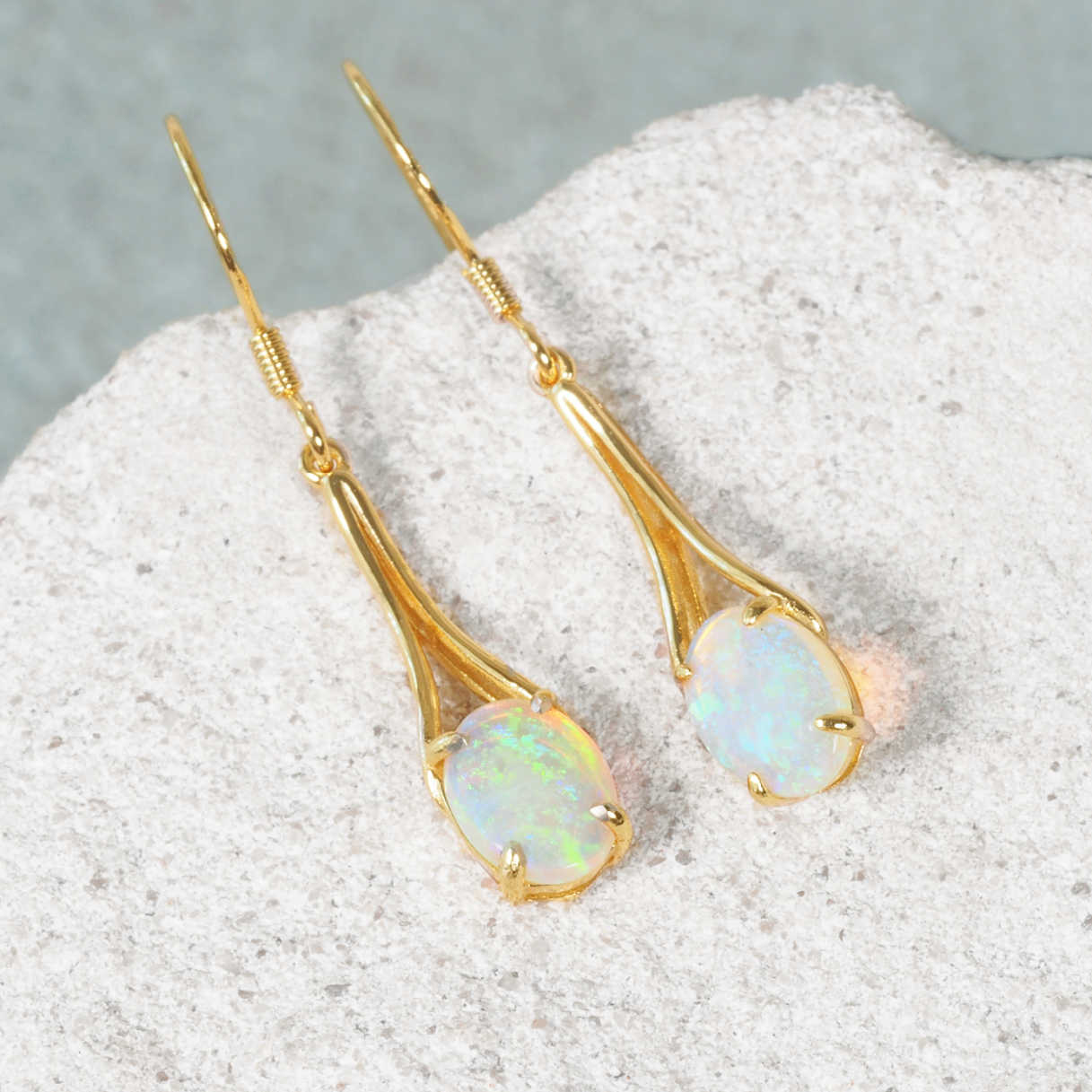 A pair of gold plated silver opal drop earrings with oval iridescent Australian opal stones, displayed on a textured light gray stone surface.