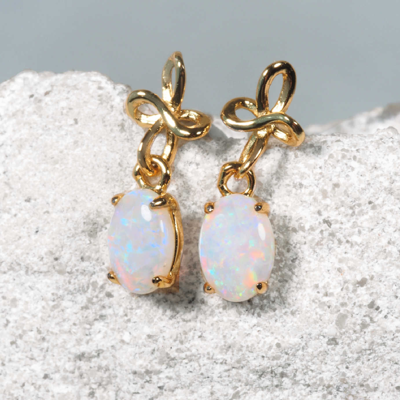 A pair of gold drop opal earrings with oval white opal stones, resting on a textured light gray rock surface.