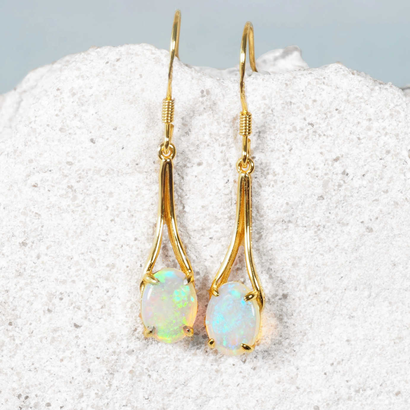 A pair of gold plated silver Australian drop opal earrings with oval green crystal opal stones, displayed on a textured white stone surface.
