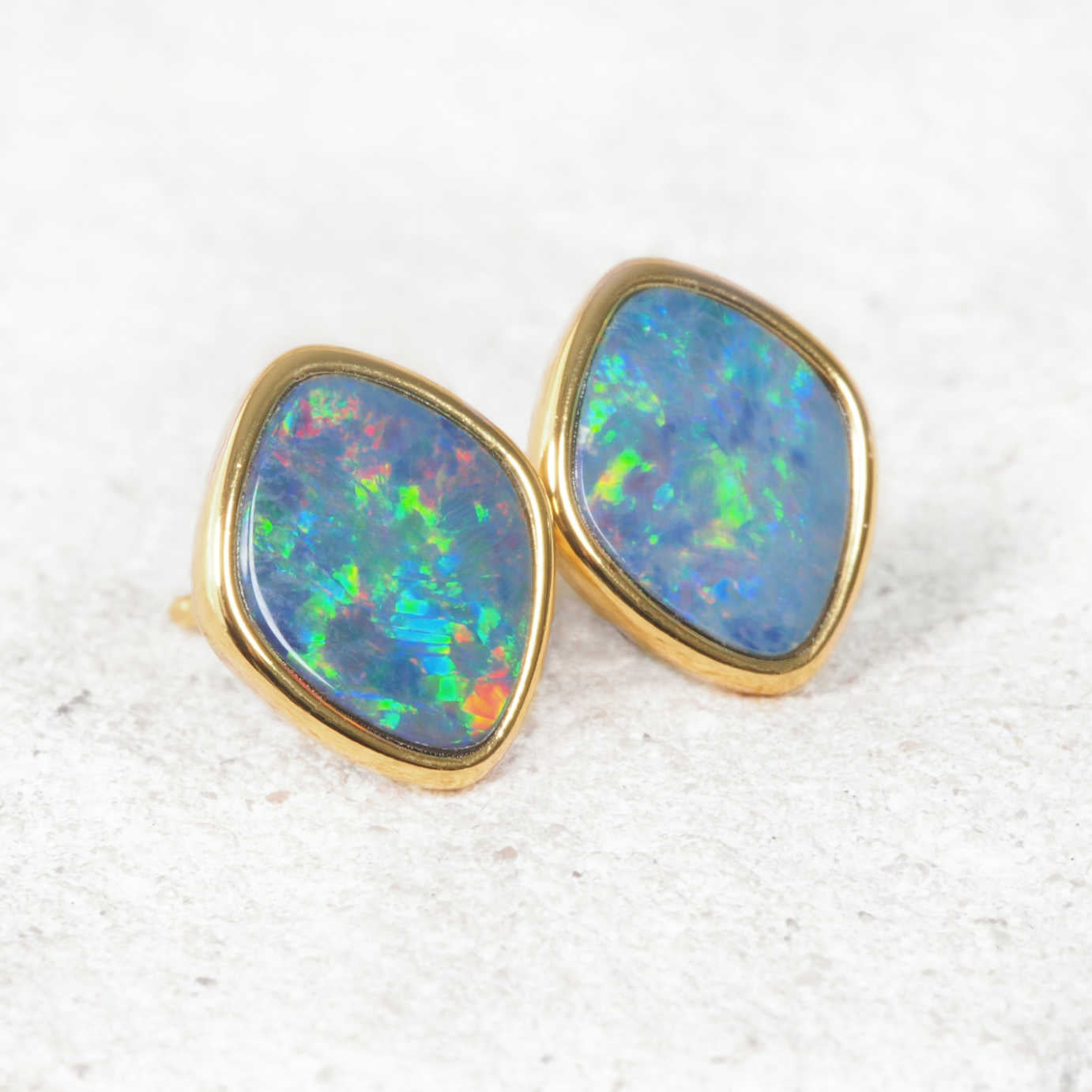 A pair of gold stud opal earrings with irregular shield-shaped blue opal inlays, displayed on a textured white surface.