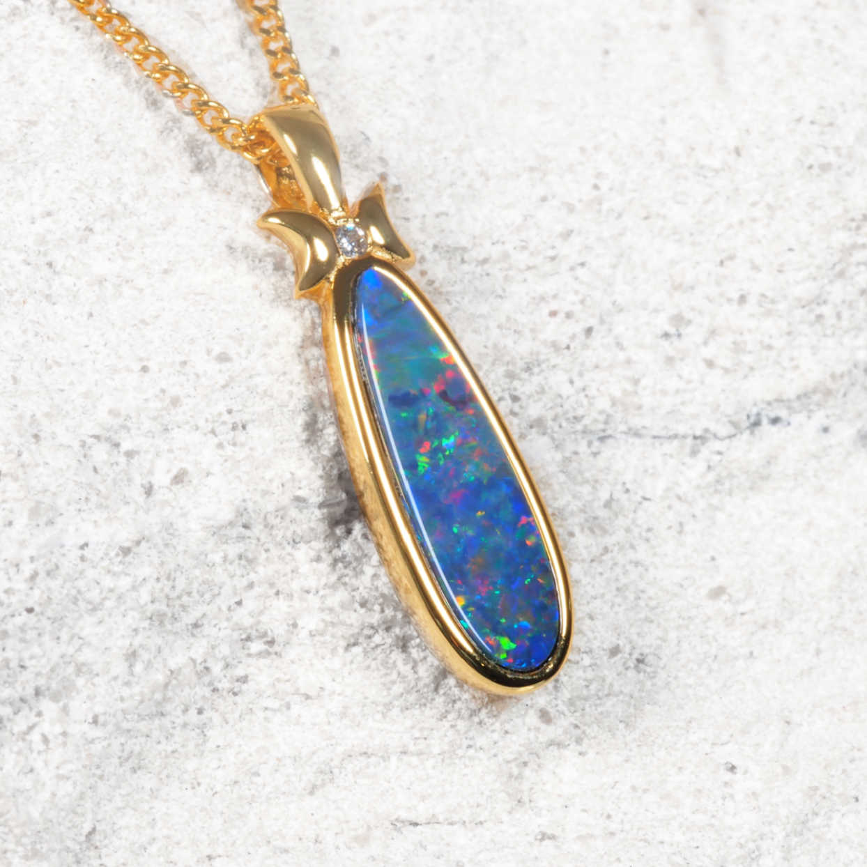 A gold plated silver opal teardrop pendant necklace featuring a vibrant blue opal with multicolored flashes, set on a textured light gray stone background.