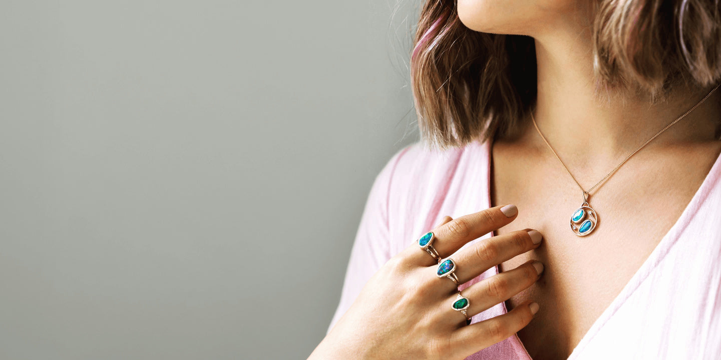 A model wears opal rings and an opal pendant while wearing a pink shirt against a grey background