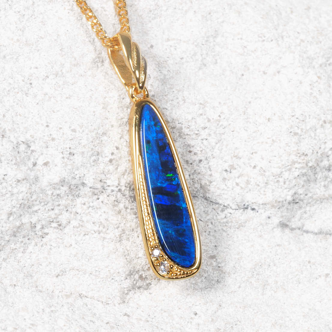 A gold plated silver Australian opal pendant necklace featuring a vibrant blue opal stone with green flashes, set in a textured gold bezel with a small clear accent stone, resting on a light gray stone surface.