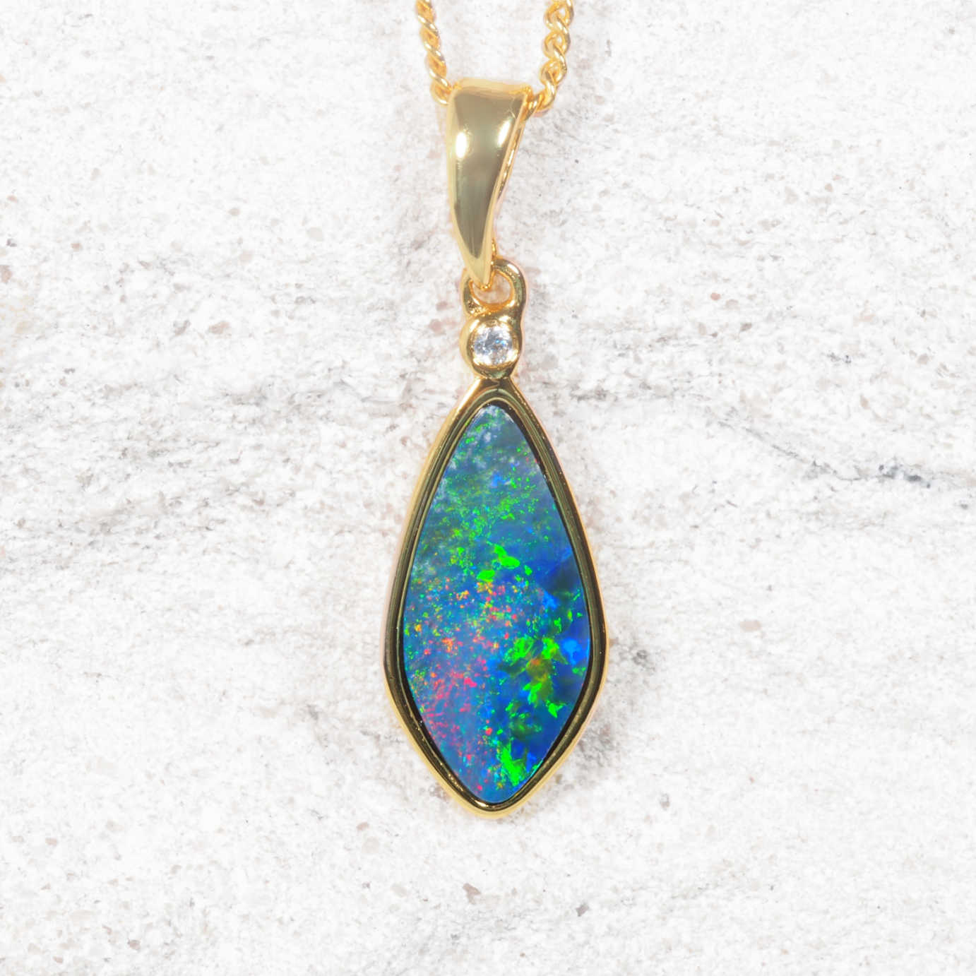 An Australian opal pendant necklace featuring a teardrop-shaped blue opal gemstone with vibrant green, pink, and orange flashes, set in a gold bezel with a small clear accent stone, resting on a textured white stone background.