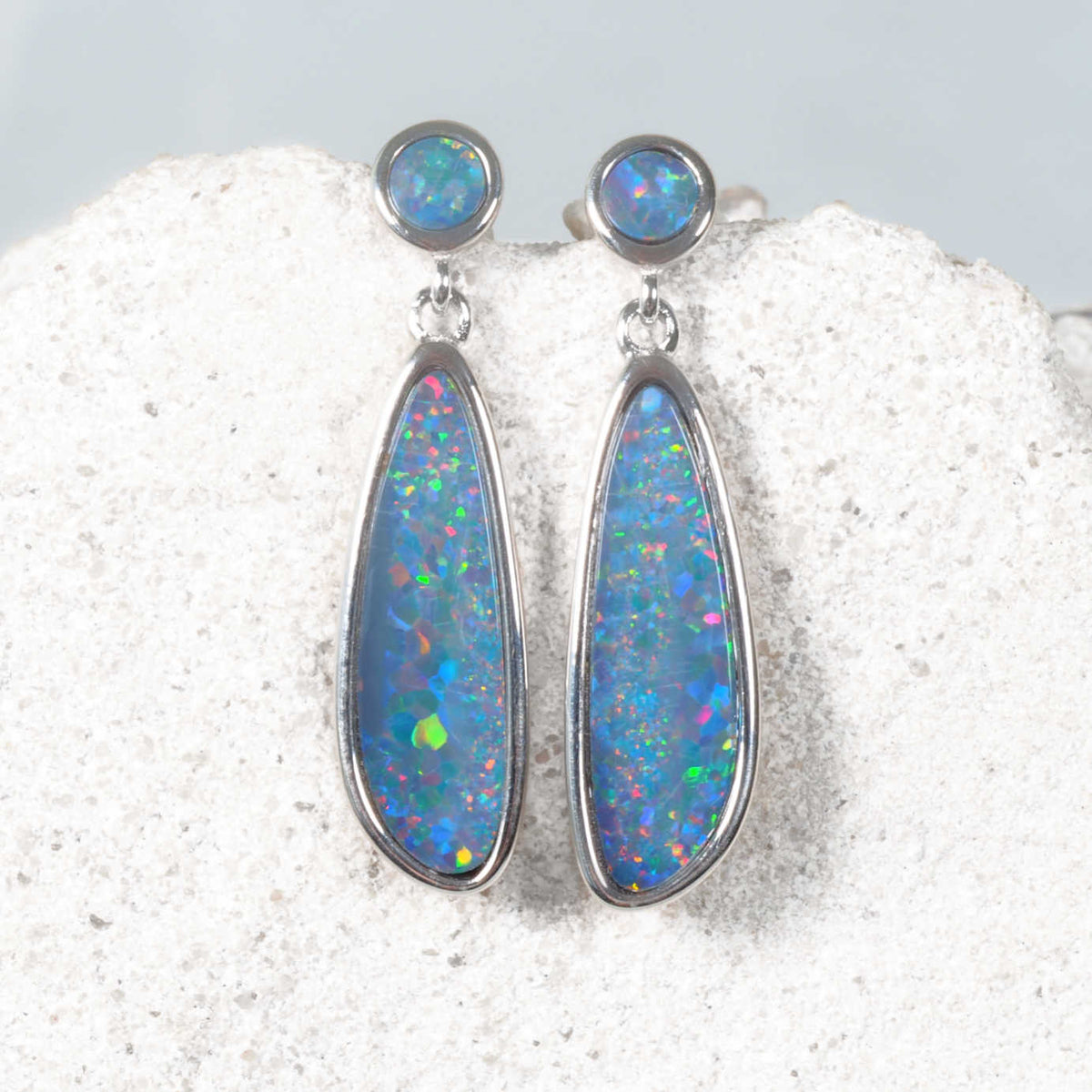 A pair of silver drop opal earrings with teardrop-shaped blue opal stones and round blue opal studs, displayed on a textured white stone surface.