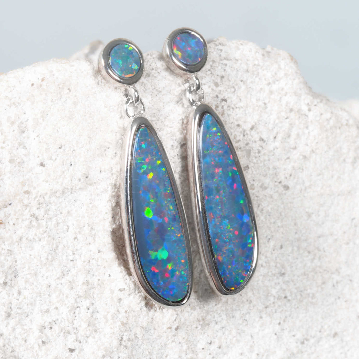 A pair of silver dangle earrings with oval blue opal stones, displayed on a textured white stone surface.