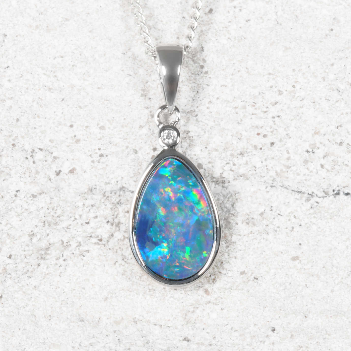 A silver teardrop opal pendant necklace featuring a vibrant blue opal stone with multicolored flashes, set in a silver bezel with a small clear accent stone.