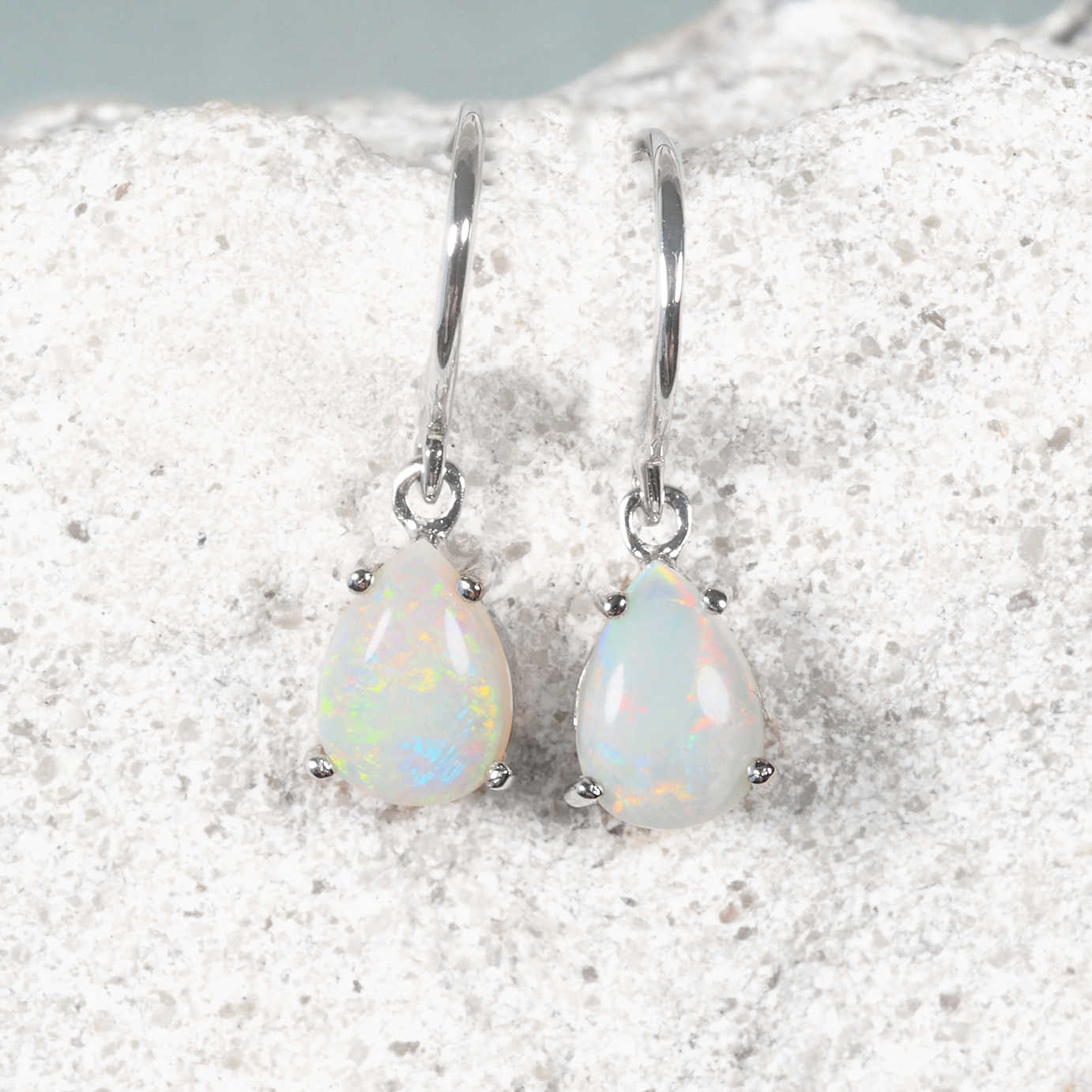 A pair of white gold teardrop dangle opal earrings with iridescent natural Australian white opal stones, resting on a textured white stone surface.