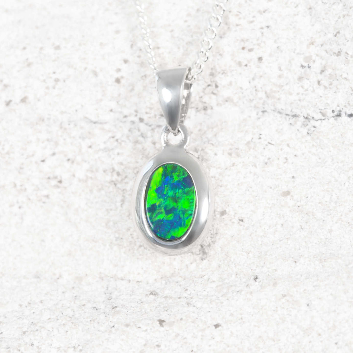 A white opal authentic oval opal pendant necklace featuring a vibrant green and blue opal stone, displayed on a textured light gray stone surface.