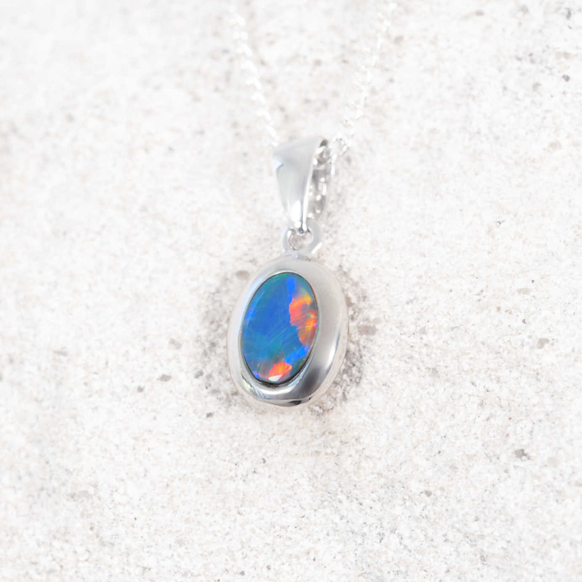 A white gold Australian opal pendant necklace featuring an oval cabochon opal with vibrant blue, orange, and green flashes, displayed on a textured light gray surface.
