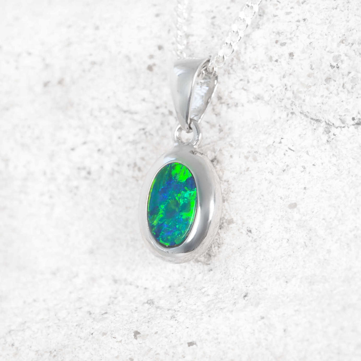 An oval pendant necklace featuring a vibrant green and blue opal gemstone, displayed on a textured light gray stone surface.