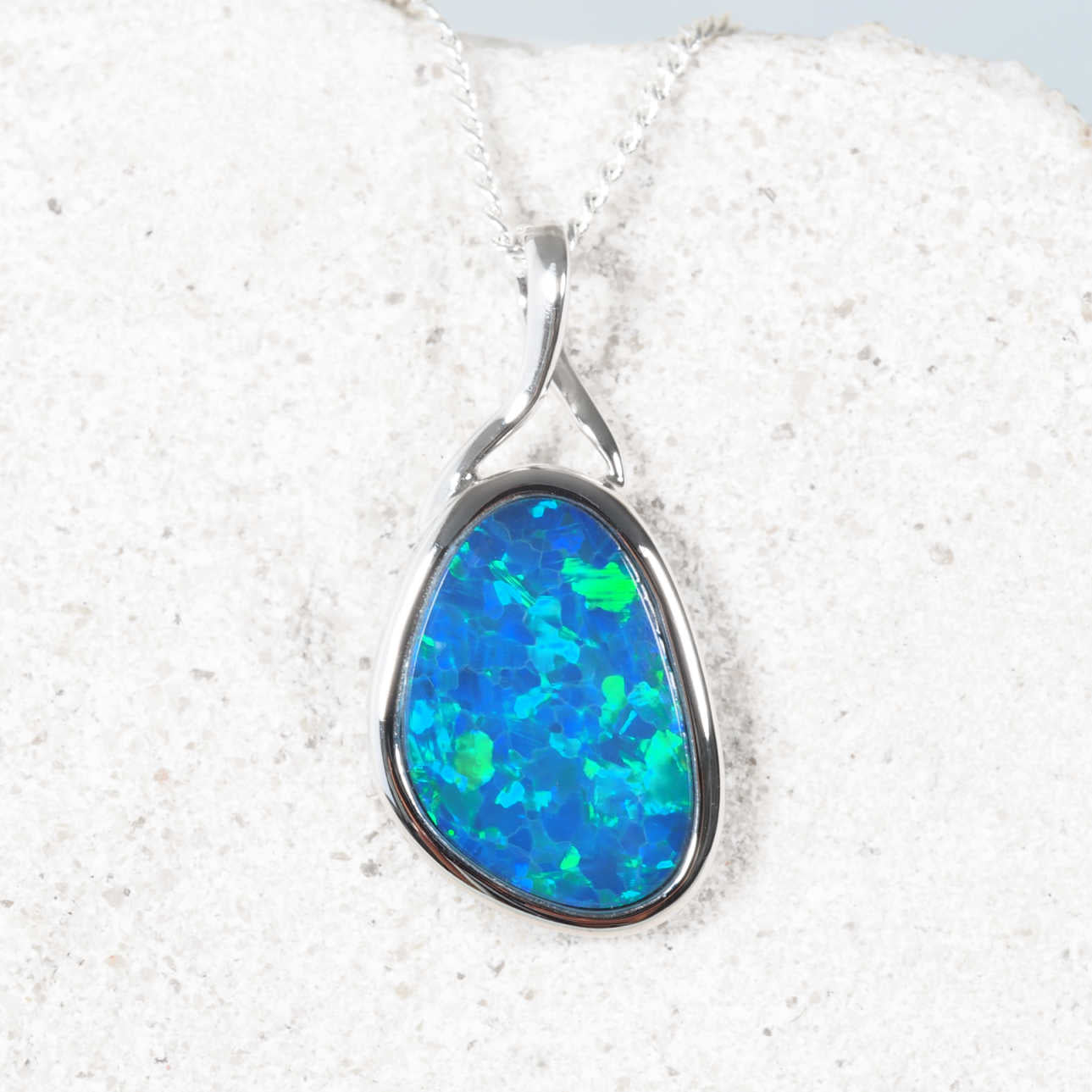 A teardrop-shaped opal pendant necklace set in 14ct white gold, displayed on a textured white stone surface. The opal features vibrant blue and green flashes.