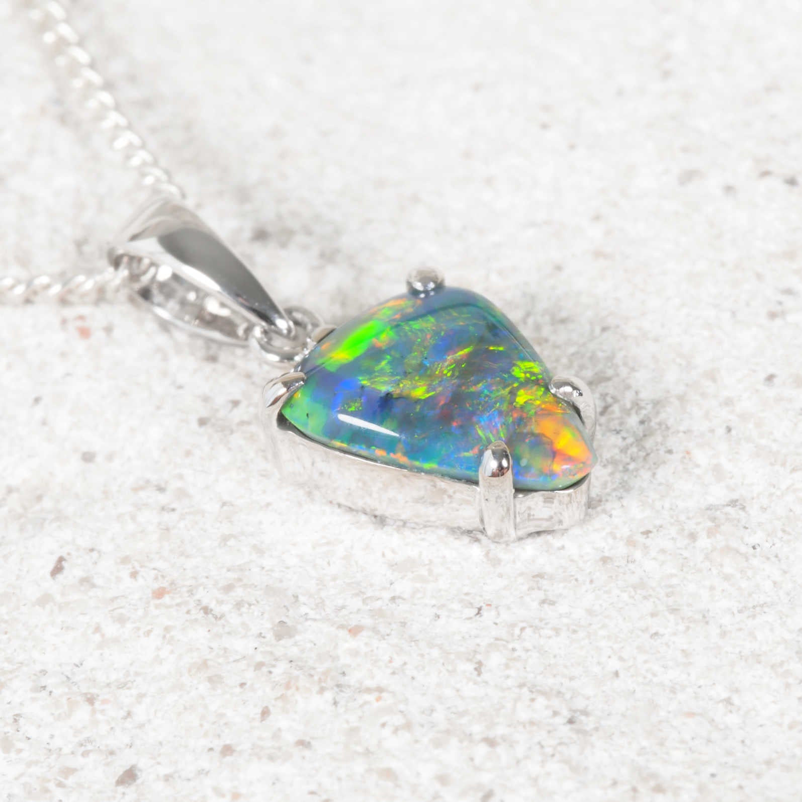 A white gold black opal pendant necklace featuring a triangular opal gemstone with vibrant green, blue, and orange play-of-color, resting on a textured light gray surface.