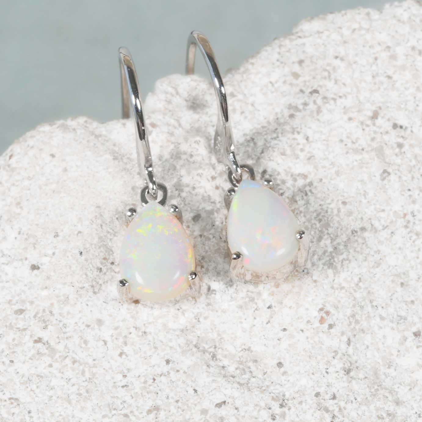 A pair of white gold opal drop earrings with teardrop-shaped Coober Pedy-sourced white opal stones, displayed on a textured white stone surface.