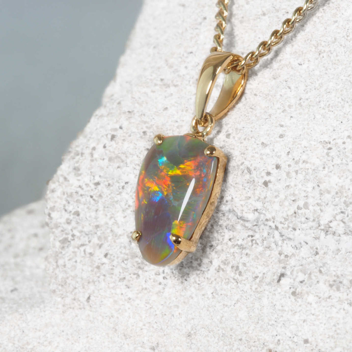 A gold opal necklace featuring an oval opal gemstone with vibrant iridescent colors, resting on a textured light gray stone surface.