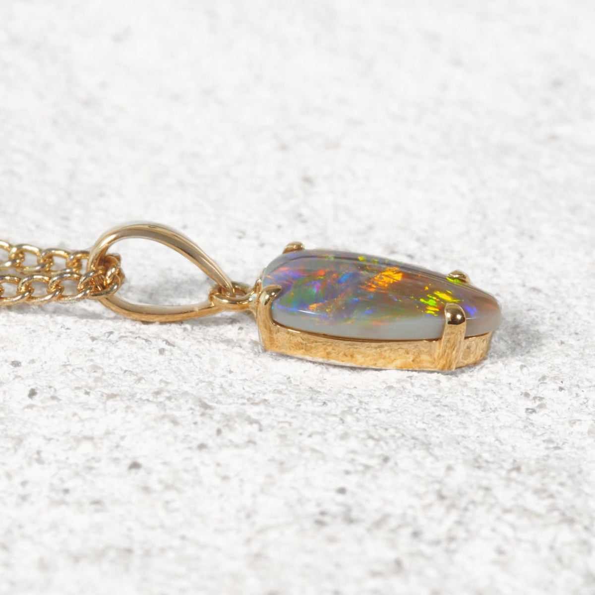 An Australian gold black opal pendant featuring a cabochon opal gemstone with vibrant iridescent colors, resting on a textured light gray surface.