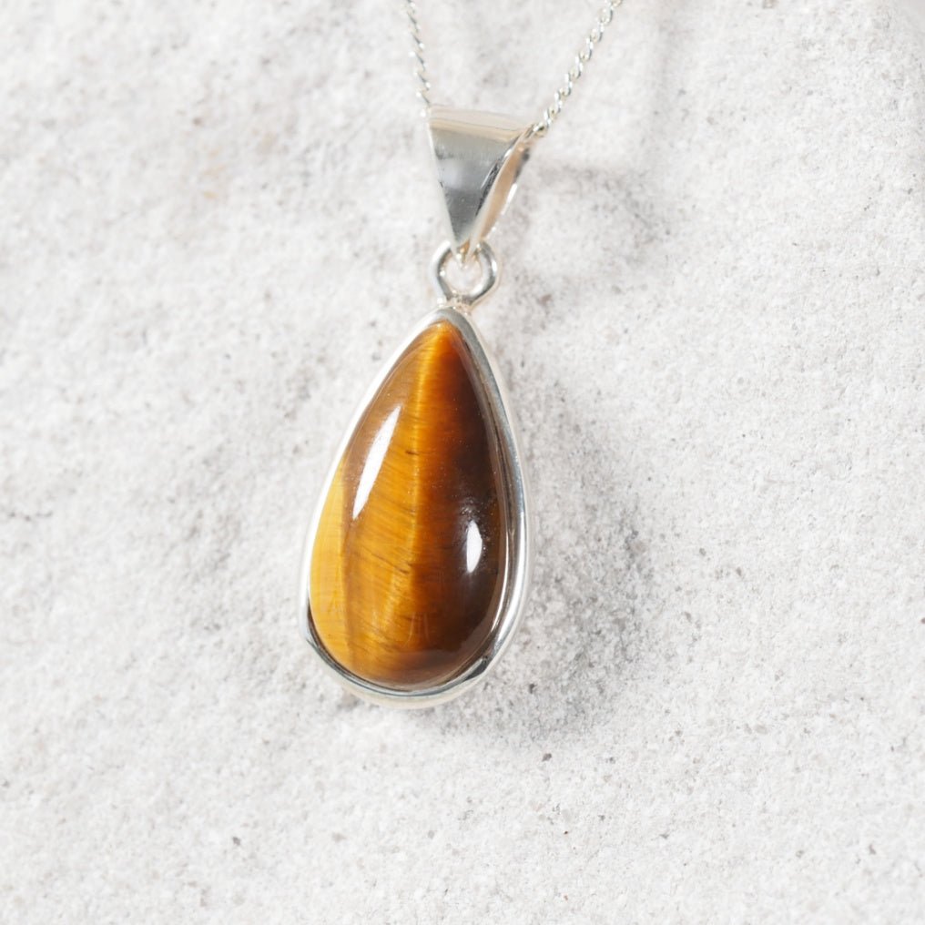 Teardrop-shaped tiger's eye stone pendant on a silver chain against a textured white background