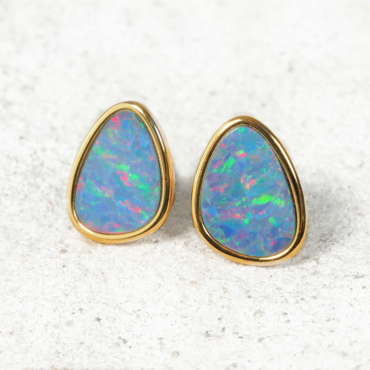 A pair of Australian gold teardrop stud opals earrings with vibrant, iridescent blue, green, and pink opal inlays, set against a light textured stone background.