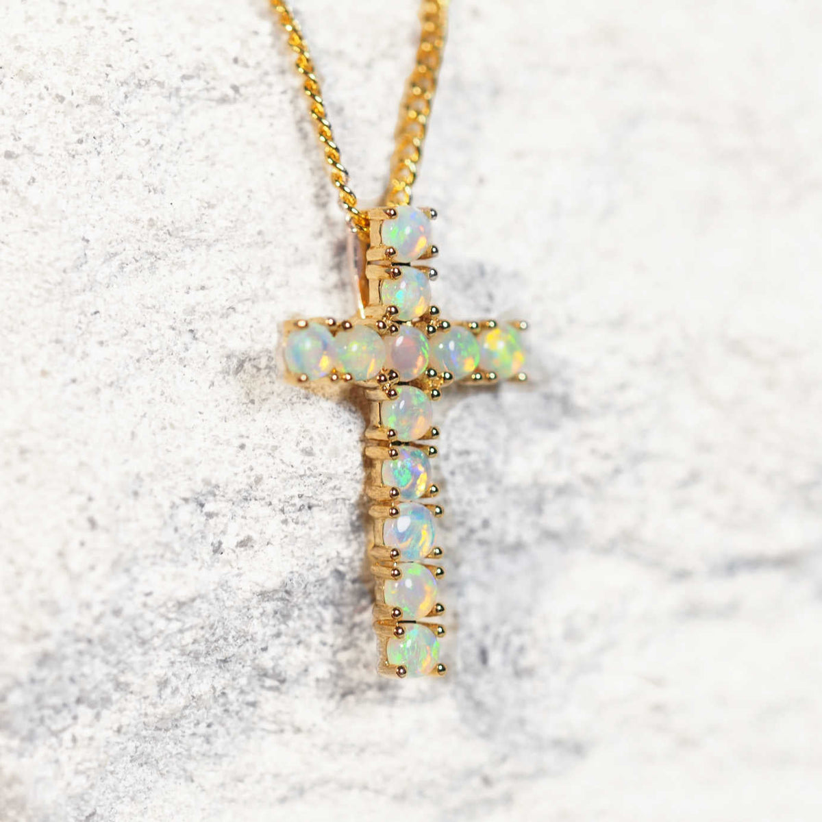 A gold cross South Australian opal pendant necklace featuring inlaid iridescent crystal opal stones, resting on a textured light gray surface.