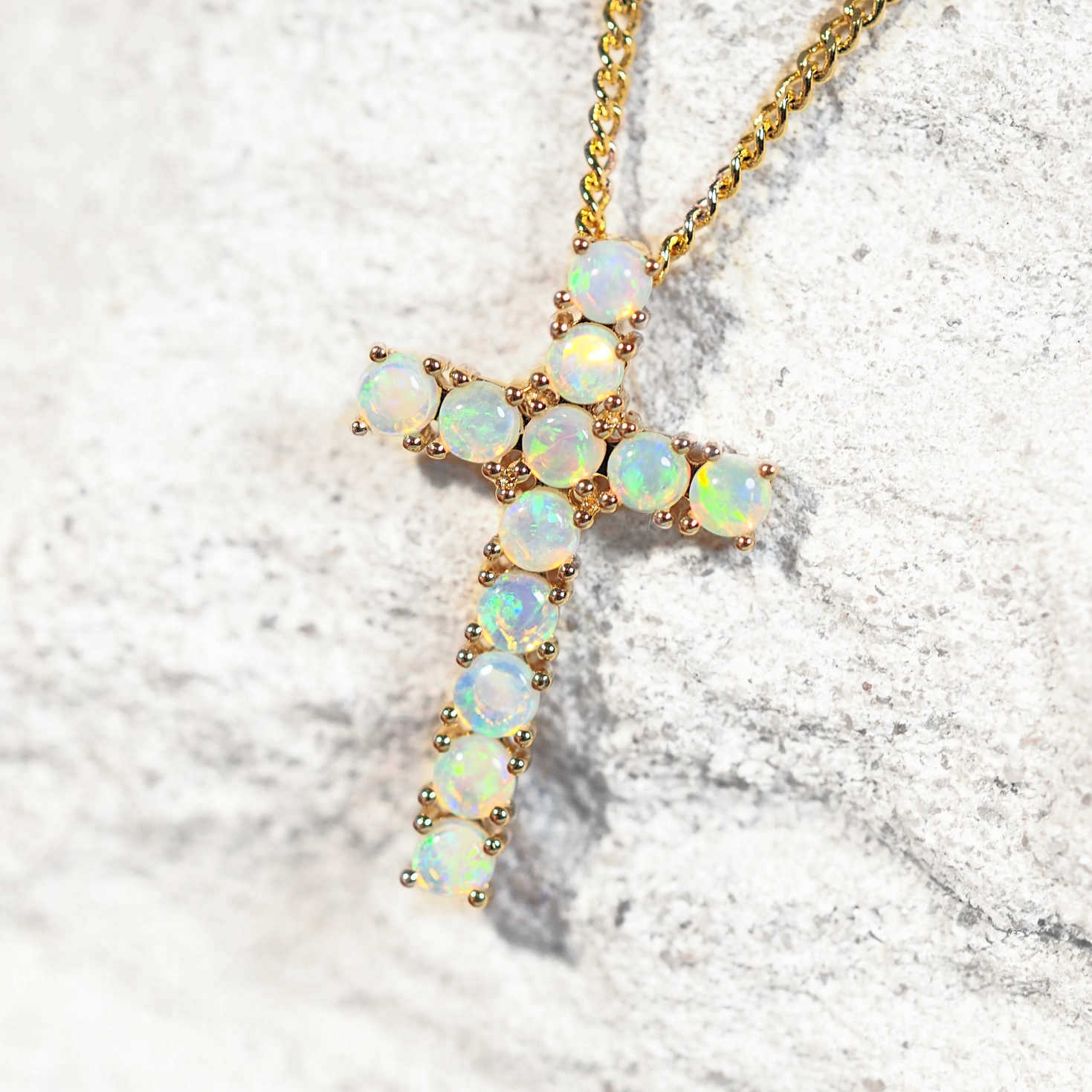A gold cross Australian opal pendant necklace adorned with round iridescent opal gemstones, resting on a textured light gray stone surface.
