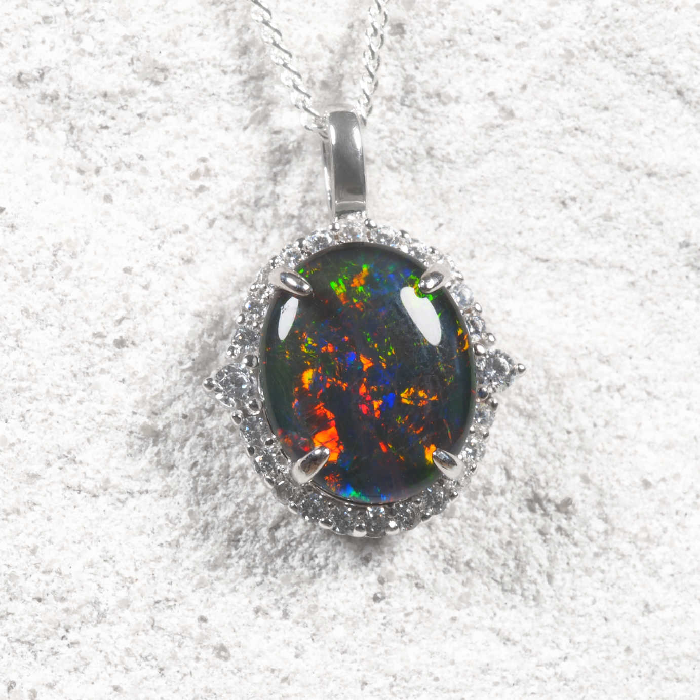A silver pendant necklace featuring a round black opal with vibrant multicolor flashes, surrounded by a halo of small clear stones, on a textured white background.