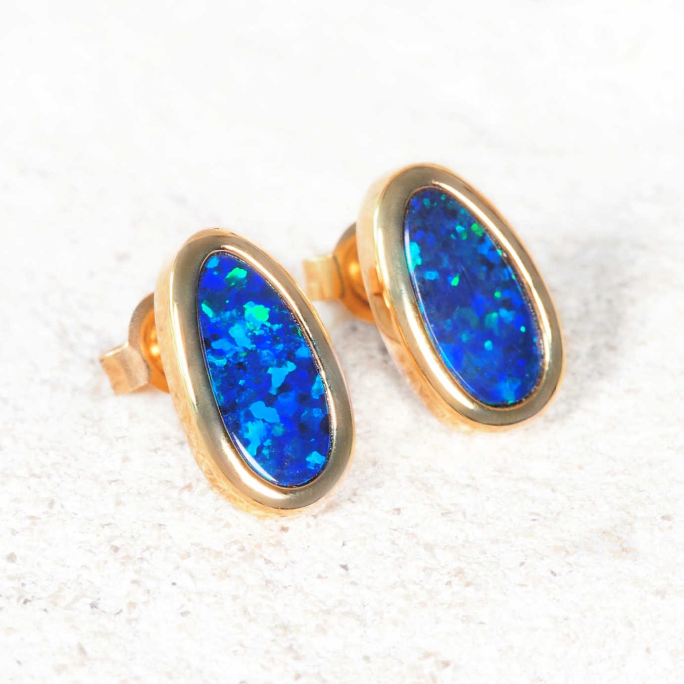 Gold-framed blue opal stud earrings on a textured white background