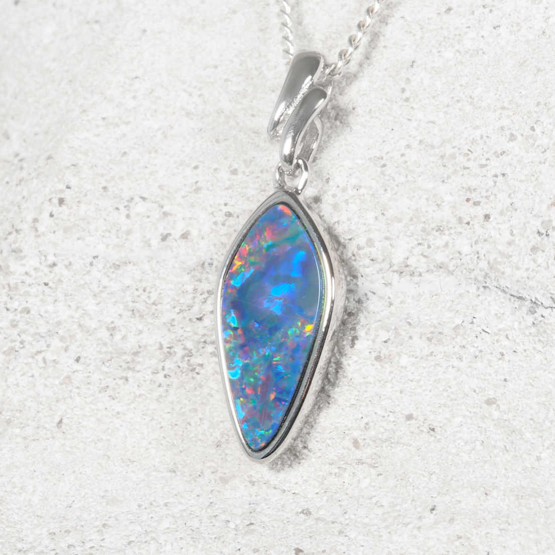 A silver opal pendant necklace featuring a teardrop-shaped opal gemstone with vibrant blue, green, and orange play-of-color, set in a polished silver bezel on a matching silver chain, displayed on a light textured stone background.