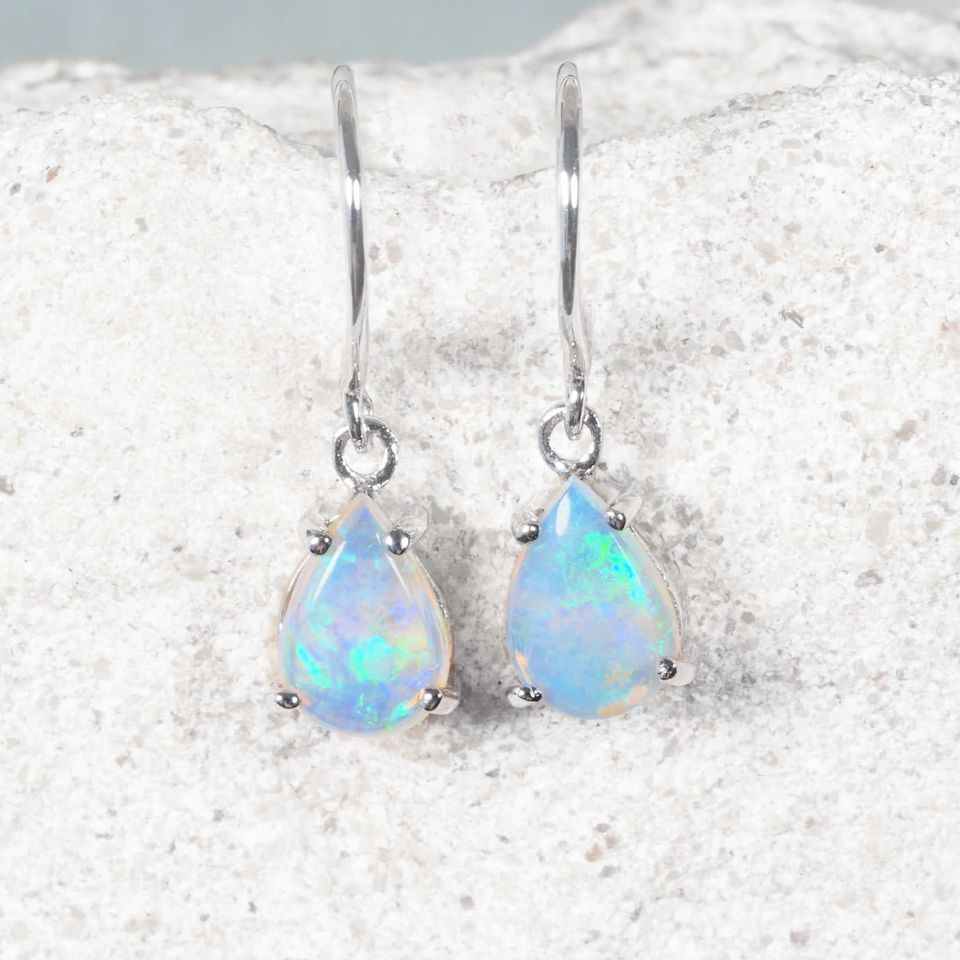 A pair of white gold authentic crystal opal drop earrings with teardrop-shaped iridescent opal stones, displayed on a textured white stone surface.