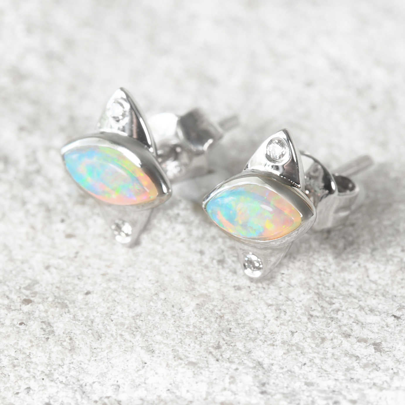A pair of authentic Australian white gold stud opal earrings shaped like cat heads, featuring iridescent marquise-cut opal gemstones and small clear crystal accents, displayed on a textured light gray surface.
