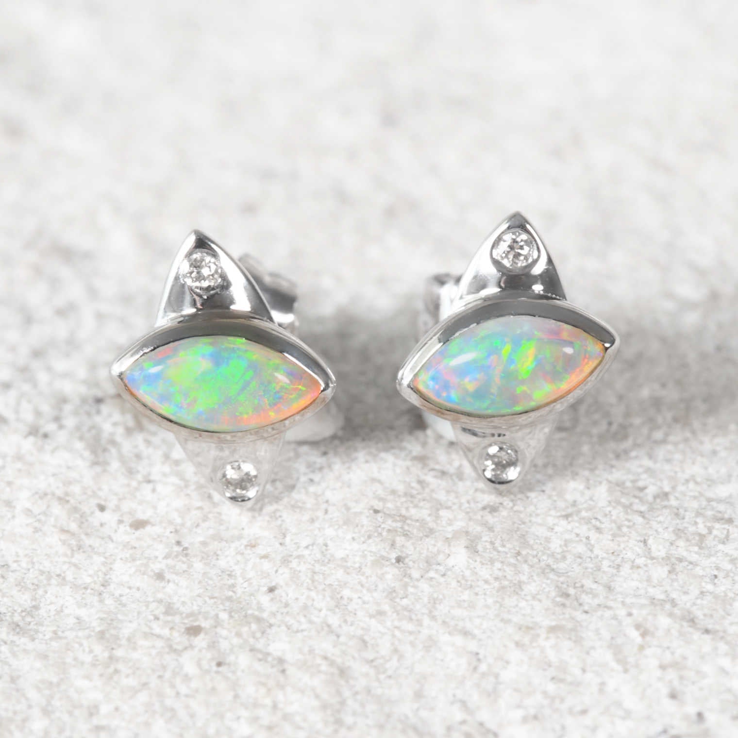 A pair of white gold opal stud earrings featuring marquise-cut iridescent opal stones with small clear accent stones, displayed on a textured light gray surface.