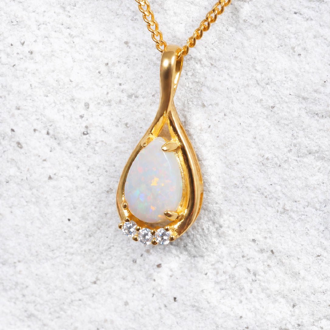 Gold plated silver teardrop pendant necklace with an iridescent white opal center and small clear accent stones, shown on a textured light gray background.