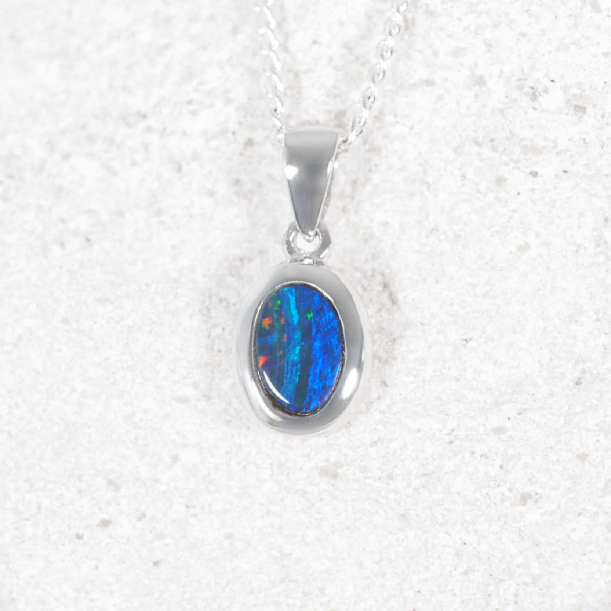 A white gold opal oval pendant necklace featuring a vibrant blue opal gemstone with flashes of orange, set on a delicate silver chain against a textured white background.